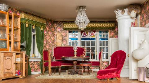 Kuva salongista, jossa tyylihuonekaluja, kristallikruunu ja plyysiverhot.