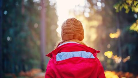 Nuori kävelee metsässä kohti auringonvaloa. Hänellä on oranssi myssy ja punainen takki.