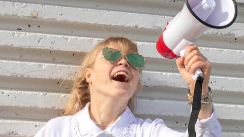 Henkilö valkoista seinää vasten megafoni kädessään ja aurinkolasit päässään