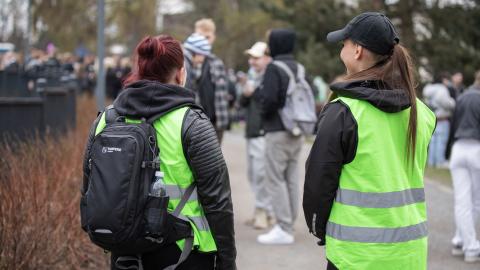 Kaksi henkilöä seisoo vihreissä huomioliiveissä puistossa, jossa on paljon ihmisiä