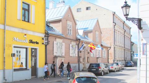 Vierekkäin kiinni toisissaan kaksi vanhaa rakennusta toinen keltainen kivitalo ja toinen vaalean ruskea vanha puutalo, joka on Tampere Maja nimeltään. Kadun varessa autoja ja ihmisiä kävelemässä kesäisessä säässä.