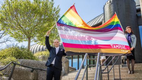Two people wave a large 13-stripe pride flag of Tampere's own on the steps of the Metso Main Library.