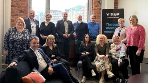 Kolmentoista hengen porukka ryhmäkuvassa. Osa istuu sohvilla, loput seisovat. Kaikki hymyilevät. 