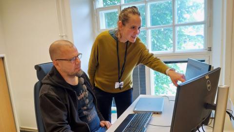 Kaksi kaupungin asiantuntijaa tutkimassa tietokoneelta Power BI -raporttia. Toinen istuu tuolilla ja toinen näyttää kädellään jotain tietokoneen ruudulla näkyvää tietoa.