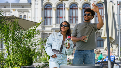 Two smiling, sunny people stand on the lush summer terrace of Tampere Central Square, with the Old City Hall (Raatihuone) in the background.