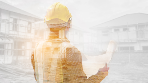 Henkilö seisoo rakennusten edessä työvaatteet päällä. Hän pitelee käsissään piirustuksia ja katsoo rakennuksia. 