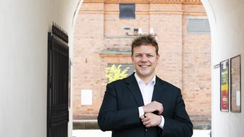 Henkilö seisoo kädet edessään ulkona valkoisten seinien välissä takanaan tiiliseinää.