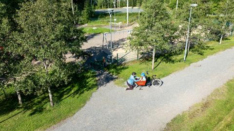 Vanhempi ja lapset leikkipuiston ulkopuolella pyörällä.