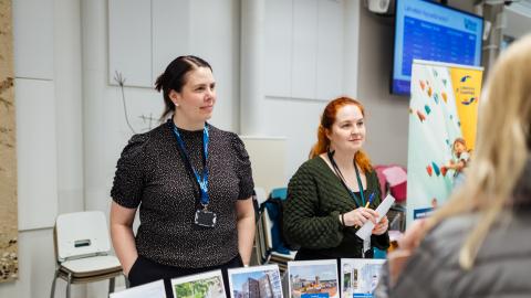 Two experts from the City of Tampere discuss with people at a residents' evening. There are several display stands on the table.