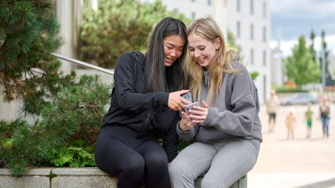 Two young women are sitting, looking happily at a phone.