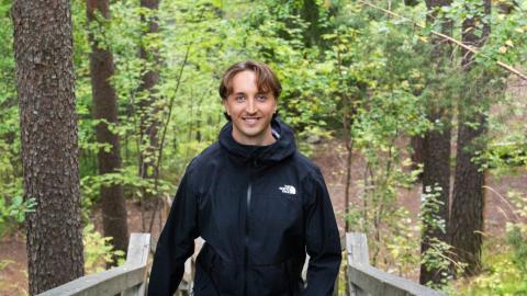 Henkilö seisoo hymyillen puuportaiden yläpäässä vehreän metsämaiseman keskellä.