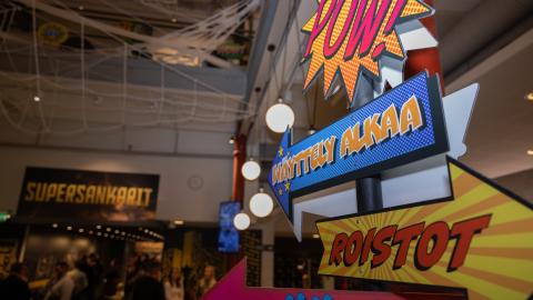 Ihmisiä Supersankarit-näyttelyn edessä, jossa myös värikkäitä kylttejä pylväässä.