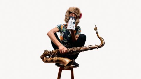 Tomoki Sanders seated on a stool holding a saxophone, wearing a floral shirt and a furry hat with flower. Their face is obscured by a small electronic device and an animal-themed mask.