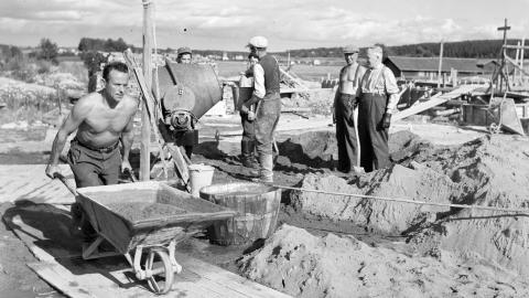 Historiallinen valokuva, jossa henkilöt seisovat rakennustyömaalla. Etualalla henkilö työntää kottikärryjä.