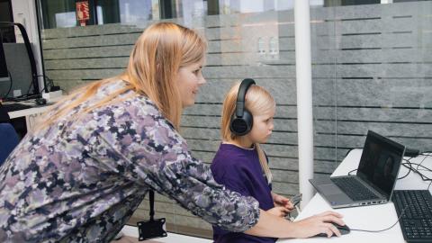 A little blonde-haired girl is sitting at a computer with headphones in her ears. Behind her is her mother, holding a computer mouse on the desk.