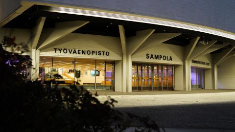 Sampola building in the dark. The wall is illuminated. There is a bush in the foreground.