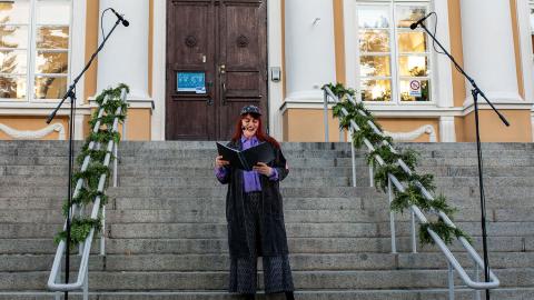 Nainen seisoo portailla ja lukee käsissään pitelemästään kansiosta runoa.