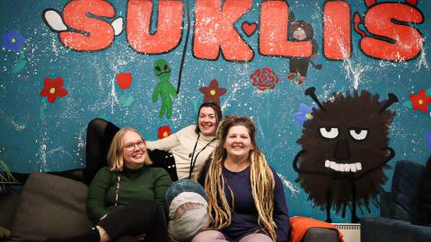 Kolme henkilöä istuu sohvalla ja hymyilee. Takana on seinä, jossa on Haisulin kuva ja jossa lukee Suklis.