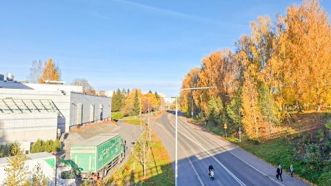 Autotie, pyörä- ja jalankulkuväylät ja kulkijoita syysväreissä olevien puiden ympäröimän, vasemmalla rakennus.