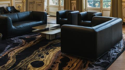 Huoneessa lattialla tumma matto, jossa on vaaleita kuvioita. Huoneessa keskellä on tumma sohvaryhmä ja nojatuolit.