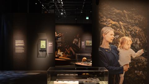 Hämärässä näyttelytilassa esineitä vitriinissä keskellä huonetta. Seinällä kuvassa äiti ja lapsi.