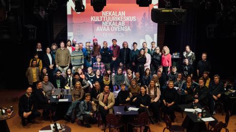 Suuri ryhmä ihmisiä poseeraa yhteiskuvassa. Taustalla valotaulu, jossa lukee "Nekalan kulttuurikeskus Nekalab avaa ovensa 1.1.2026".