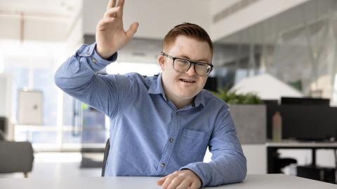 Kauluspaitaan pukeutunut henkilö, jolla on Downin oireyhtymä, nostaa toista kättään.