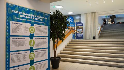 Seinällä juliste, jossa on paljon tekstiä. Taustalla huonekasvi ja portaikko.