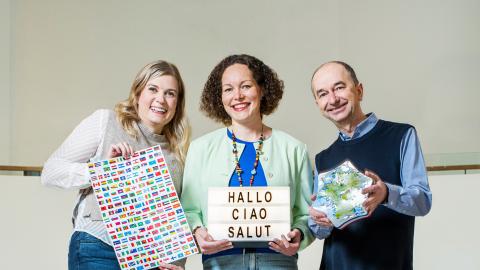 Three people are standing side by side. They are holding language learning materials, such as flags and a blackboard.