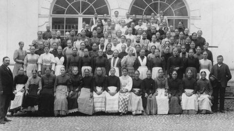 Mustavalkoisessa valokuvassa Finlaysonin tehtaan henkilökuntaa ryhmäkuvassa. Naisilla on pitkät esiliinat. Taustalla seinässä kaksi kaari-ikkunaa.