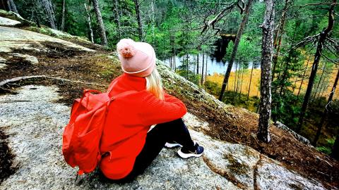 Punatakkinen tyttö reppu selässään istuu kalliolla ja katselee maisemaa, jossa näkyy metsää ja pieni järvi.