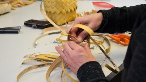 A person is weaving birch bark strips at a table. In the background, there is a birch bark box.