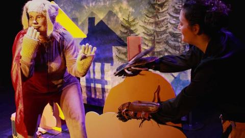 A person dressed in a dog costume holds "paw" in front of their mouth and smiles. In the background are spruce trees and a silhouette of a house.