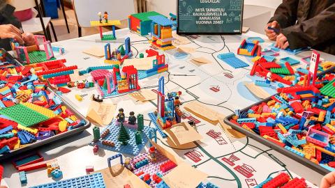 On a large table, a pile of colourful Lego blocks is being used by people to build the neighbourhood of their dreams on a map.