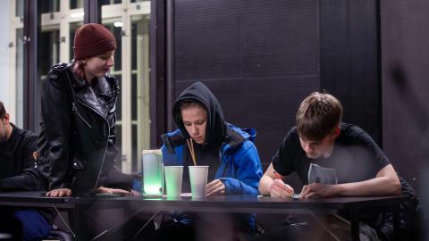 Kaksi nuorta istuu pöydän ääressä kirjoittamassa ja yksi nuori istuu pöydällä ja hymyilee