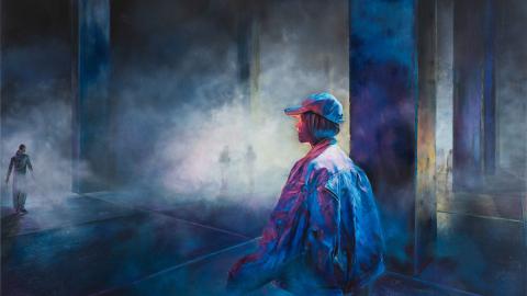 Nuori lippispäinen mies seisoo hämyisessä, tyhjässä tehdashallissa. 