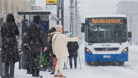Talvisella pysäkillä nainen pysäyttää bussin, jossa lukee: Matkalla kanssasi.