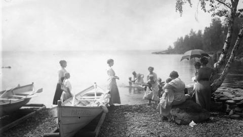 An old photograph showing people on the shore of a lake. Boats in the foreground and trees on the edge.