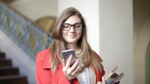 Pitkähiuksinen nuori henkilö katselee kännykkää. Toisessa kädessä on kirjoja. Taustalla julkisen tilan portaikko ja pylväs.