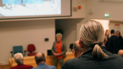 People are sitting in a lecture hall. In the background, there is a screen and a lecturer.