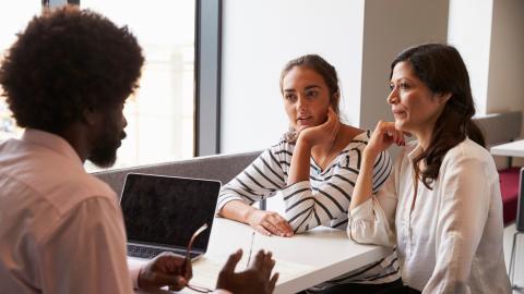 A teenager and a mother are sitting at a table. On the other side of the table is a teacher with a computer.