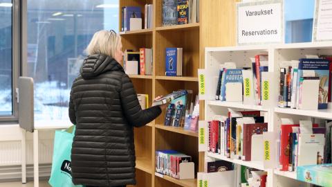 The customer is examining the back cover of a book and standing in front of the reservation shelf at Vuores Library.