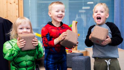 Three small children look at the camera, laughing, holding small brown stuffed box in their hands.