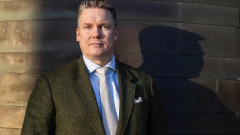 A person is standing in a dark suit in front of a dark wall. A dark shadow is visible beside them.