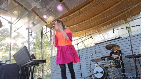 Pinkki-oranssiin mekkoon pukeutunut laulaja esiintyy ja mustiin pukeutunut rumpali soittaa taustalla.