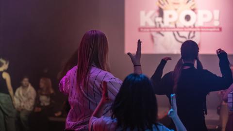 Ryhmä nuoria tanssii salissa, jonka seinällä on heijastettu kuva, jonka päällä lukee k-pop