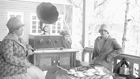 Mustavalkoisessa valokuvassa kaksi naista istuu ulkona kuuntelemassa vanhanaikaista radiota.