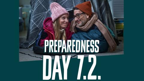 A warmly dressed woman and man smile at each other in a picture with the words "Preparedness Day 7.2." written on it.