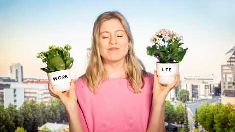 The person has their eyes closed and is balancing flower pots in both hands. One pot says "work" and the other says "life." In the background is the cityscape of Tampere.