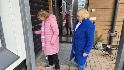 A woman with an intellectual disability, dressed in a pink padded jacket, is unlocking the front door of her home. Her mother, dressed in blue, is standing behind her.